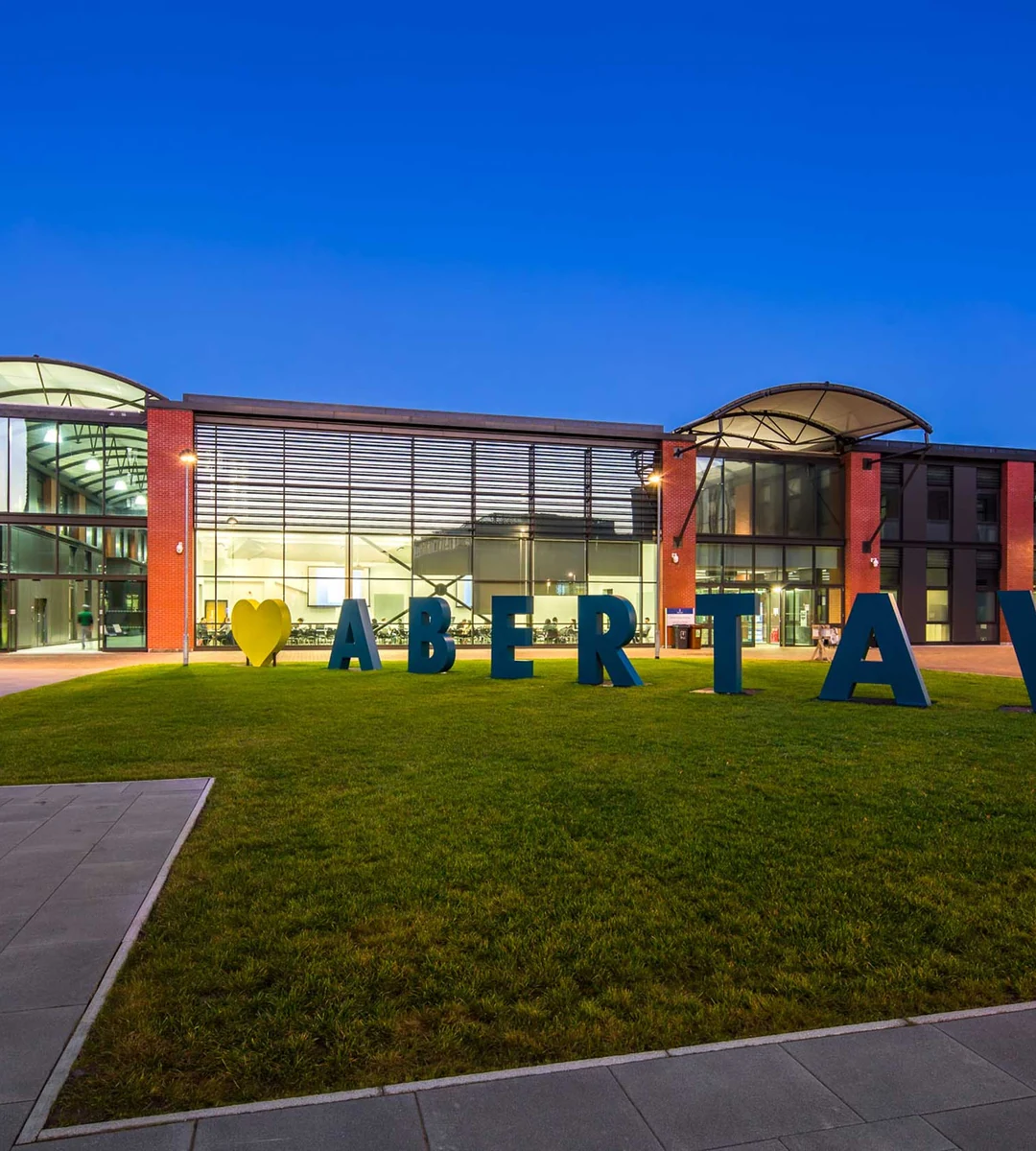 Корпус Bay Campus Swansea University вечером и объёмные буквы ABERTAWE на газоне